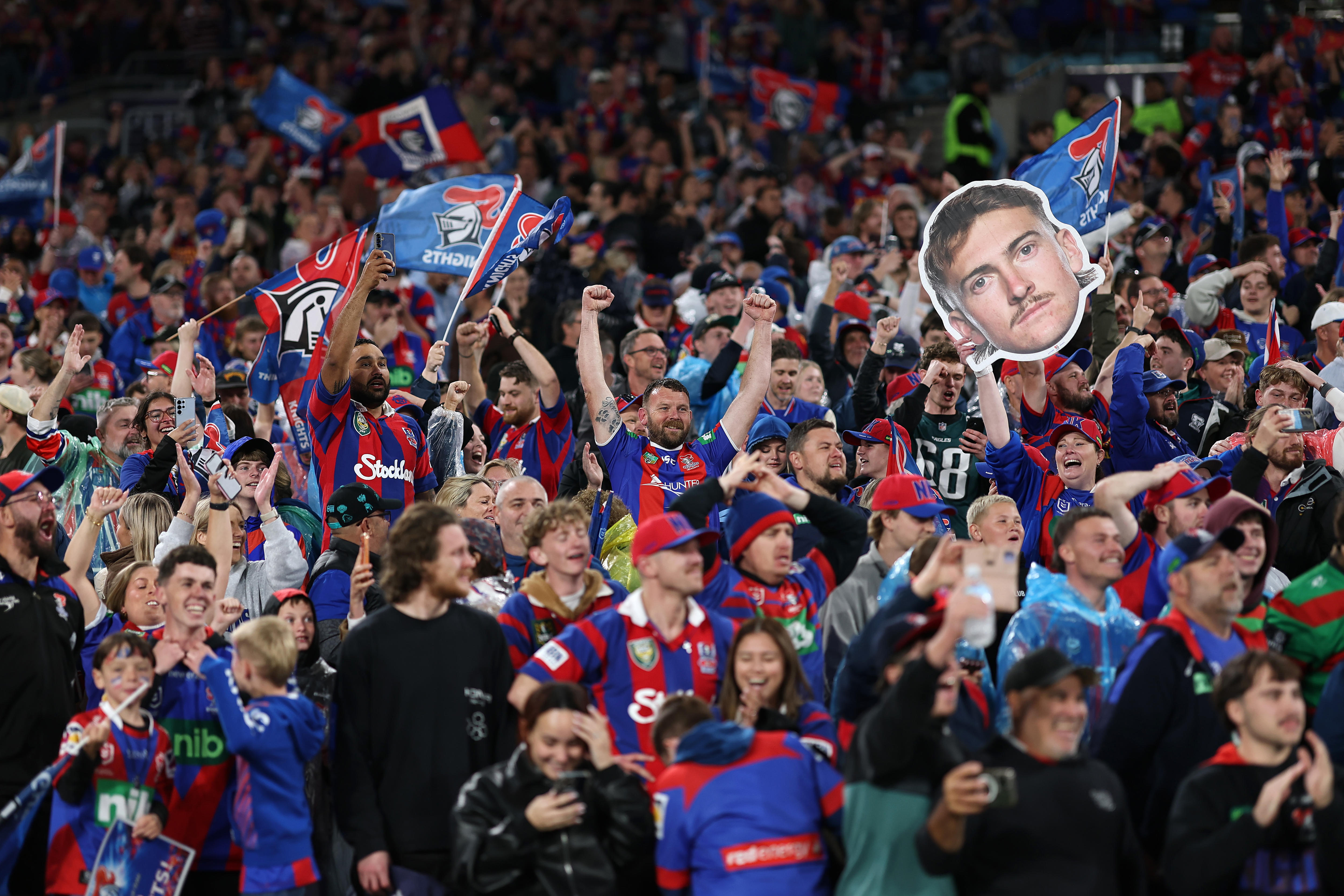 A group of rugby league fans in red and blue cheer and hold up banners