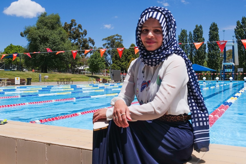 A woman in blue skirt and headscarf sits on the edge of an outdoor 50 metre pool.