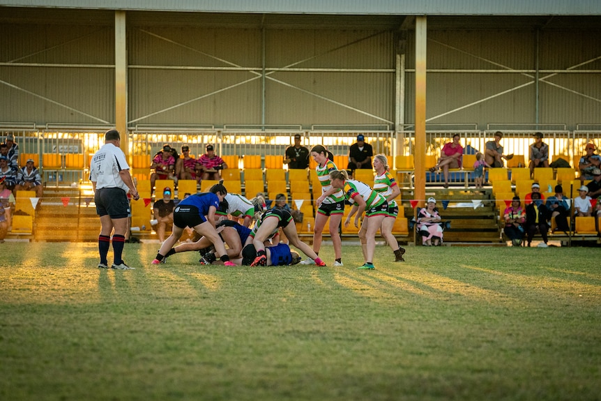 A womens sports team dressed in green and white play a game of footy on an open field in front of an audience