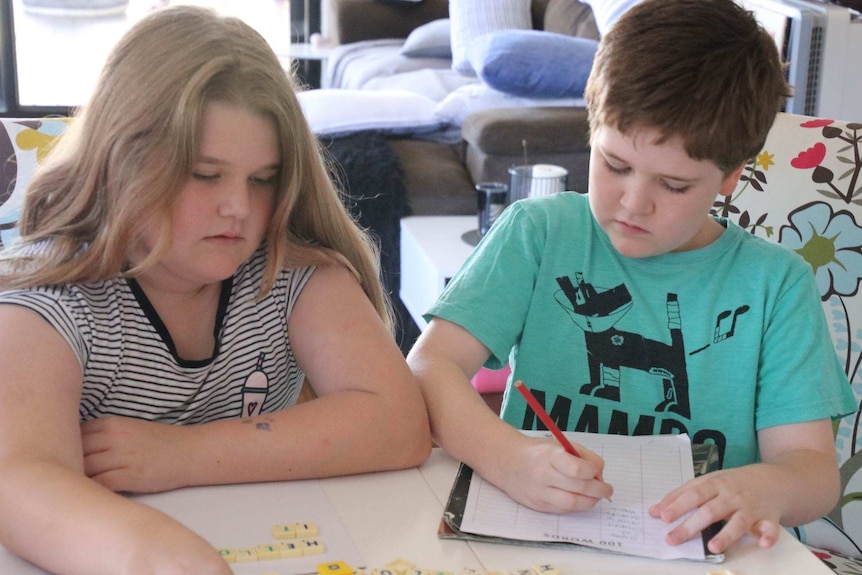 A young girl sits at a table making words with Scrabble letters as her brother sits next to her writing.