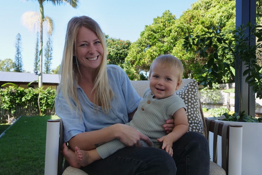 A mother and son sit on a chair, the sunny garden in the background, they are both grinning at the camera