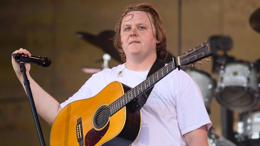 Lewis Capaldi stands in a white shirt with an acoustic guitar, a hand on a microphone stand and looks wistfully past the camera