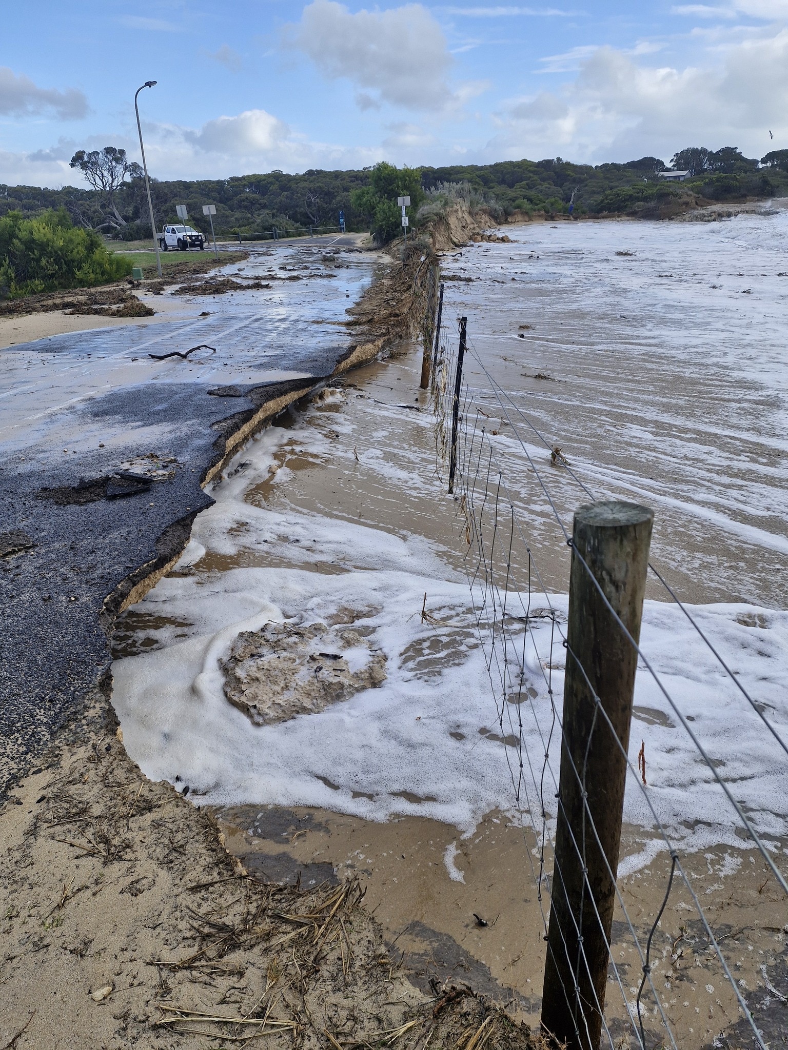 Uma estrada danificada pela água com ondas passando por baixo de uma cerca e atingindo-a.