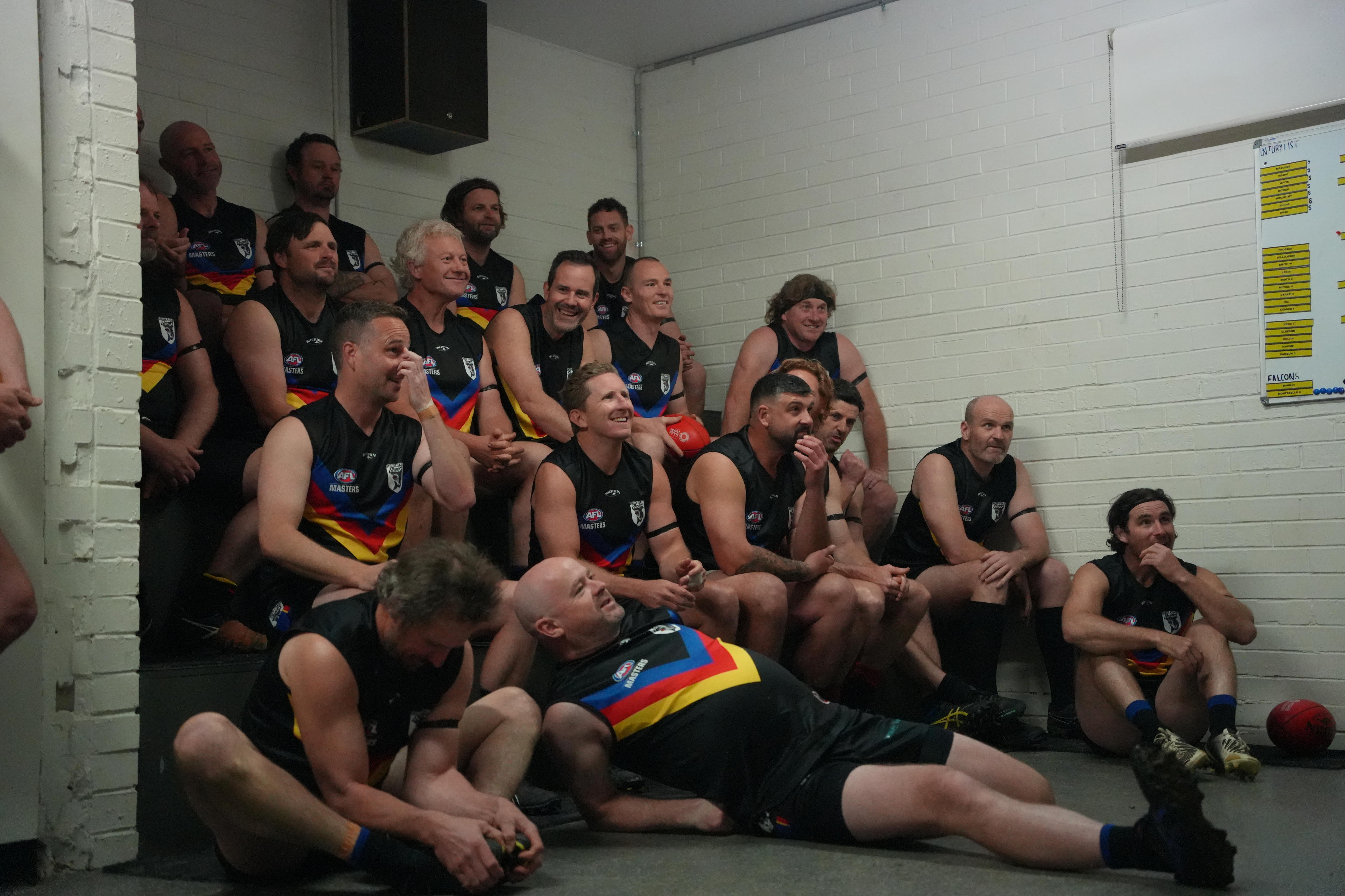 The whole South Coast Sharks football team, about 20 men, sit on benches in the changerooms while wearing their football uniform
