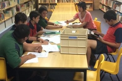 Students sit around a desk while a teach talks to them.