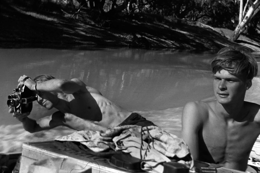 A black and white photo of two young shirtless men near a river, one looks through binoculars.