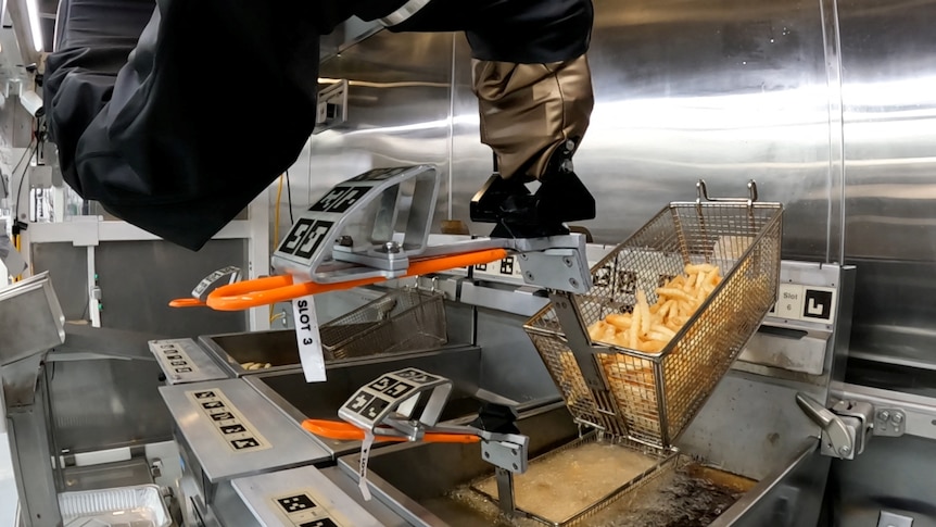 A robotic arm holds a tray of hot chips over a vat of oil in a makeshift fast-food kitchen.
