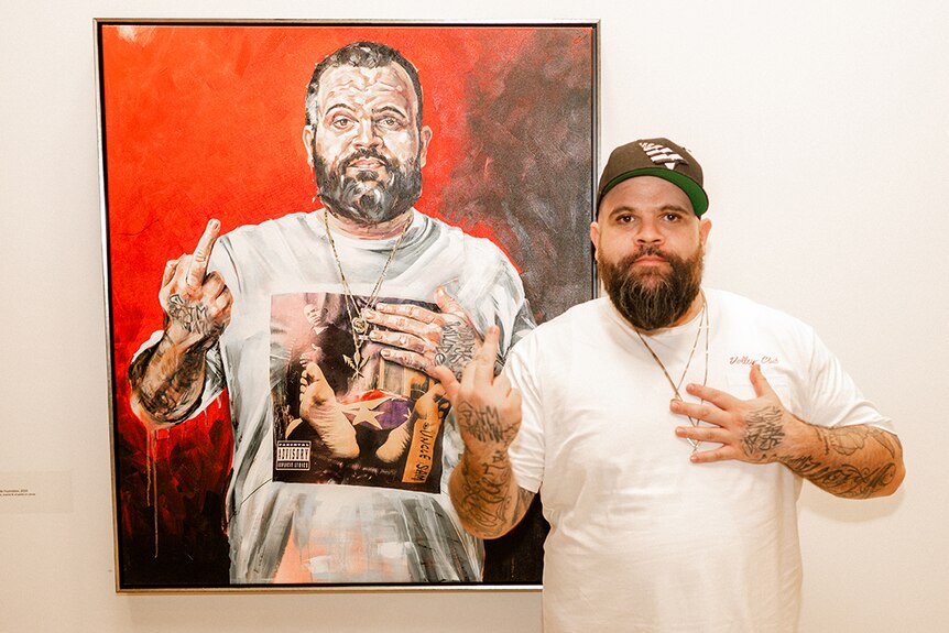 Briggs poses next to a painting of himself with his hand on his heart and middle finger raised