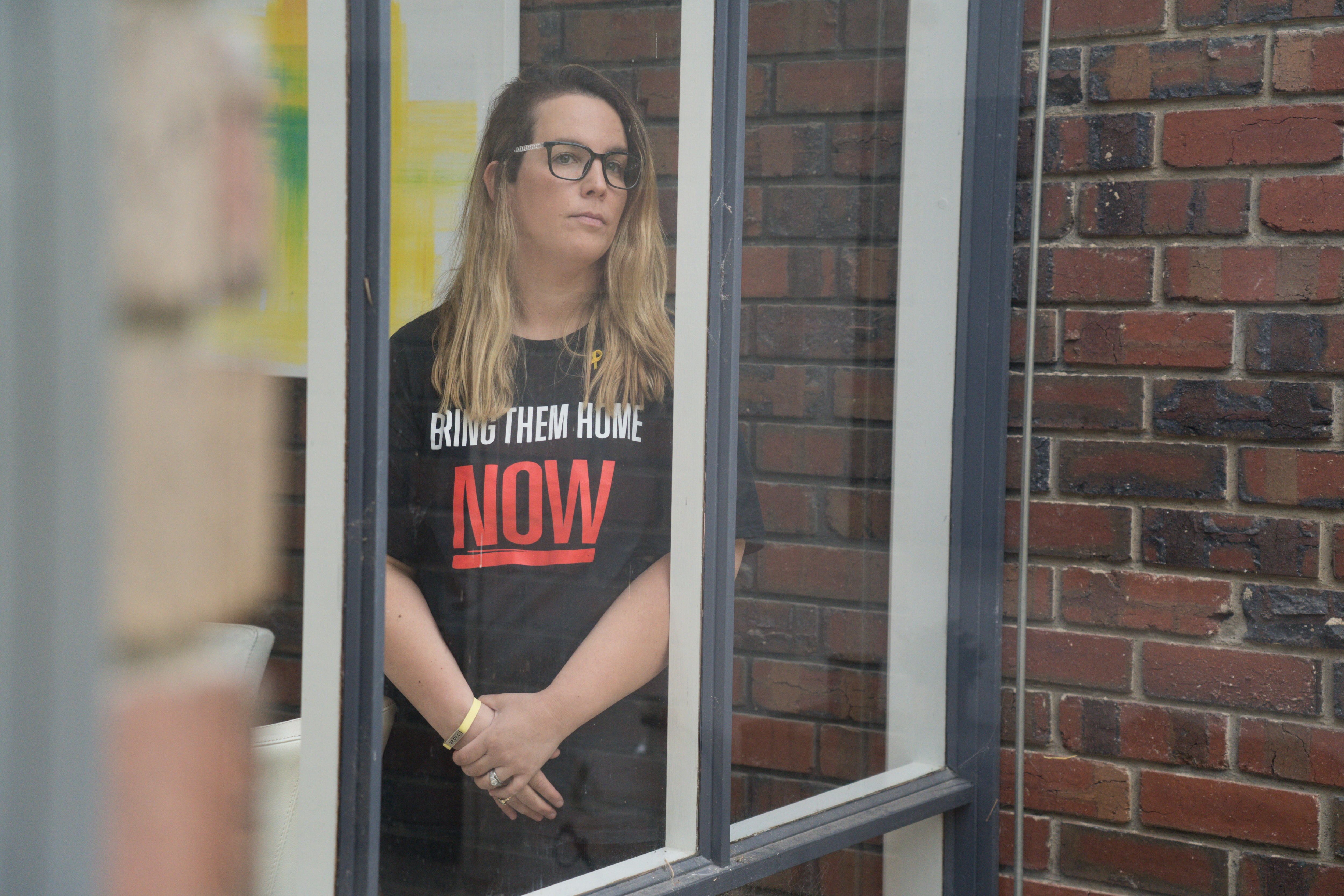 A woman wearing a shirt reading 'Bring Them Home Now' looking out a window