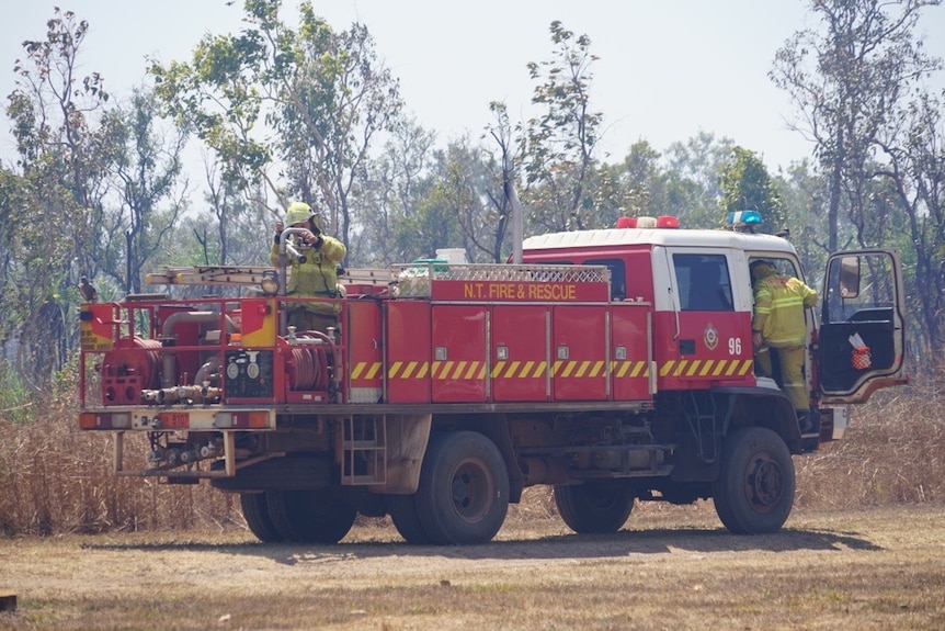An NT Fire and Rescue fire truck is parked next to bushy scrub with two firefighters.