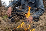 Male seated in dry grass with small fire in foreground