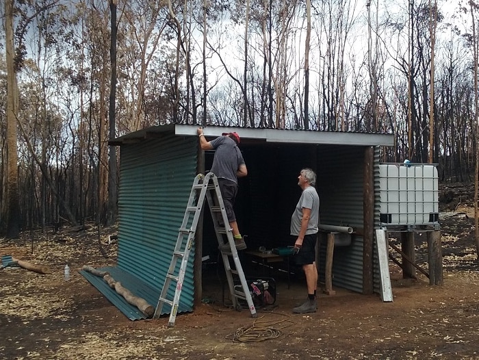 A shed stands in front of burnt out trees. Two men stand in the shed, one on a ladder near the roof.