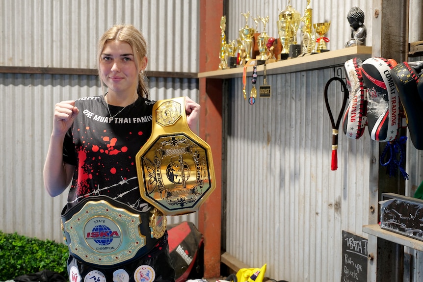 A young woman holds a championship belt buckle and wears another, she holds one hand in a fist near her head. 