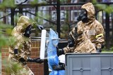 Military personnel wearing masks wrap a spade in a plastic sheet.