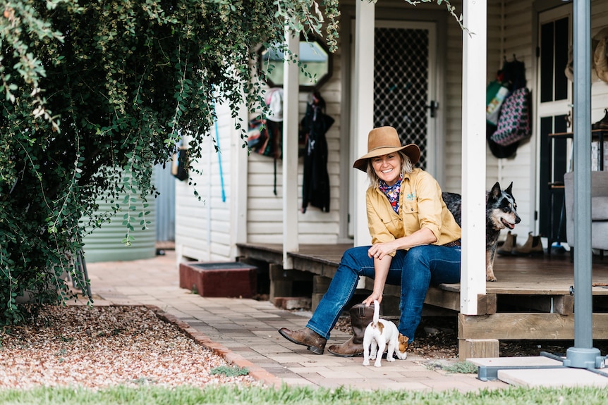 Shanna Whan sits on her back deck in yellow workshirt and wide brim hat, smiling at camera, with her two dogs sitting with her. 