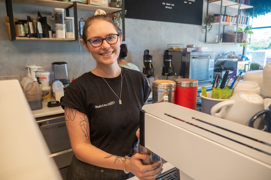 A young woman smiles behind a coffee machine.