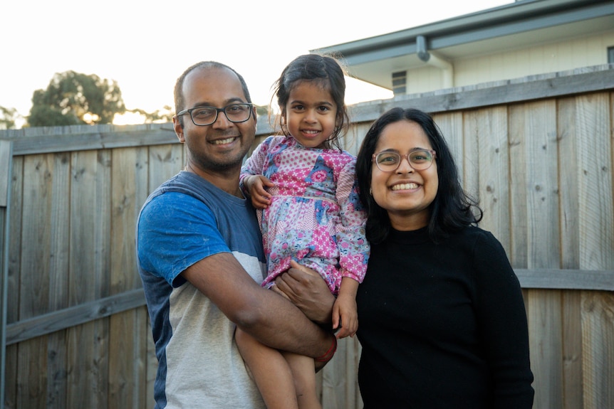A man and a woman stand in front of a timber fence, holding their three-year-old daughter between them