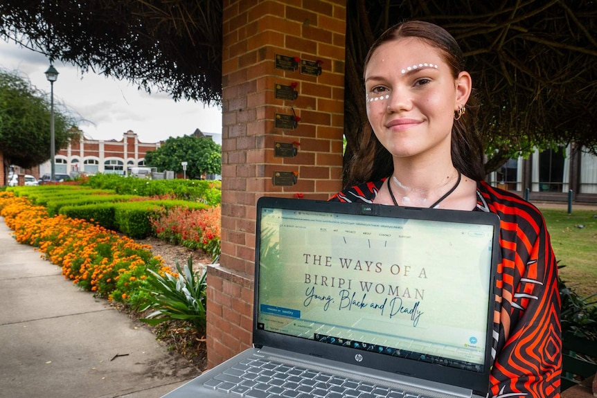 A young indigenous woman holding a laptop to camera that features her blog and the words 'The ways of a Biriipi woman'