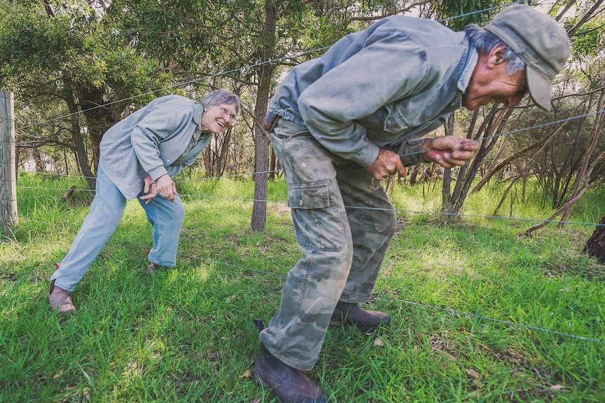 An older man and woman smile as they climb through a gap in a fence, surrounded by bush.