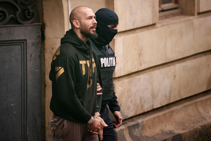 Andrew Tate is escorted in handcuffs by a balaclava-clad police officer from a court building