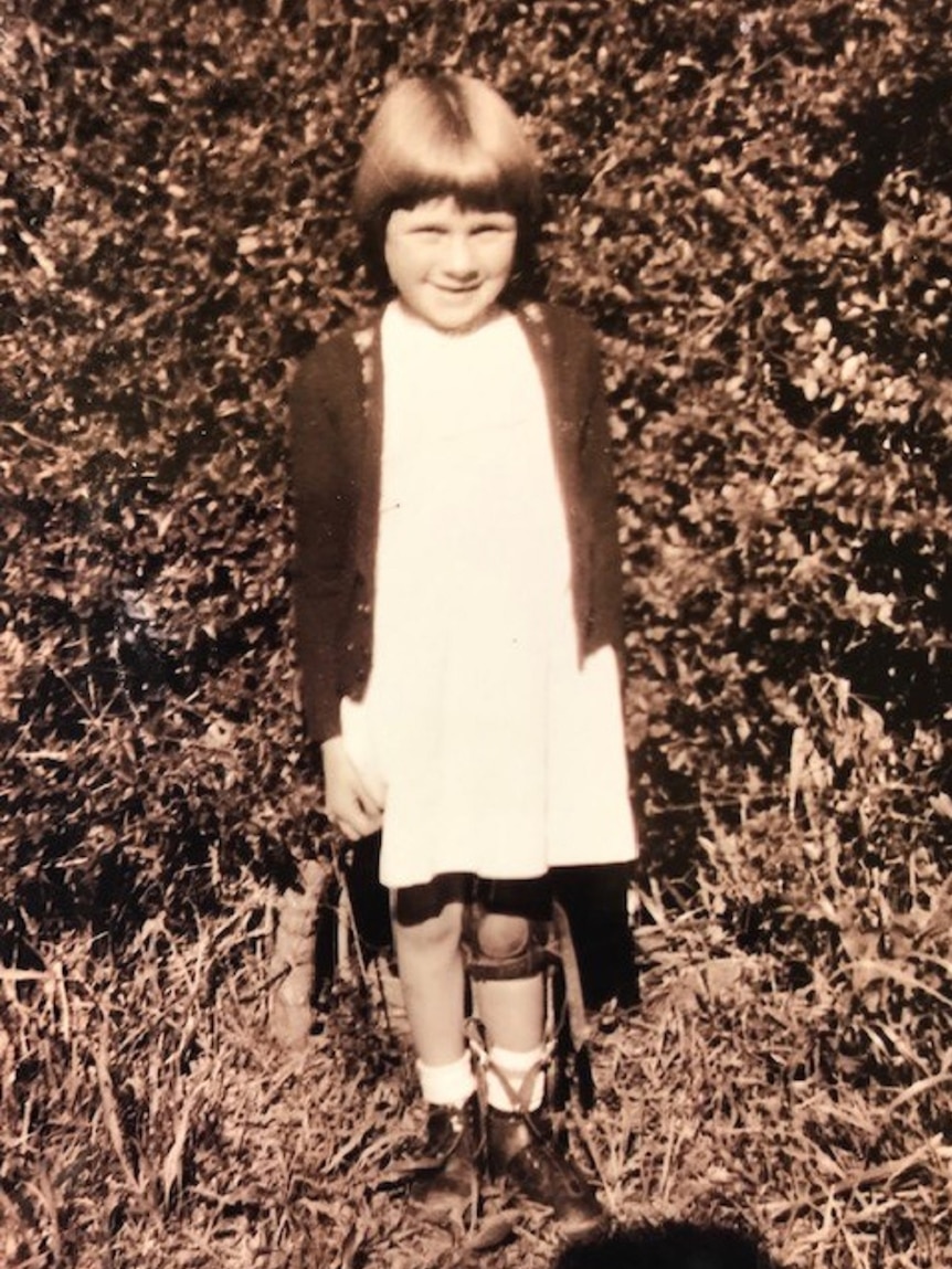 A young girl wearing a caliper on her left leg stands in front of garden hedge