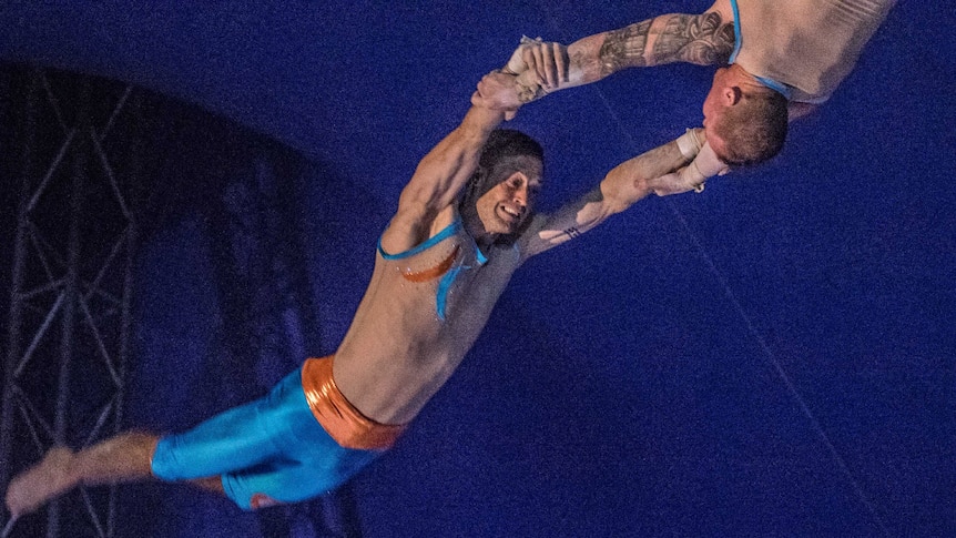 A male trapeze artist swings through the air held by another man.