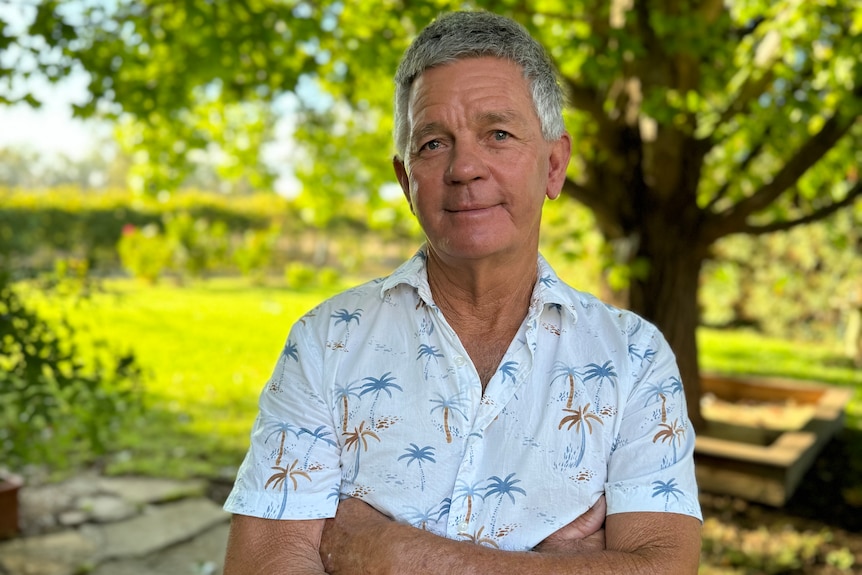 Grey-haired, fair-skinned man, Peter, under shaded trees with his arms crossed.