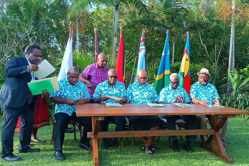 Pacific leaders wearing matching blue Hawaiian shirts sit at table with flags in the background. 
