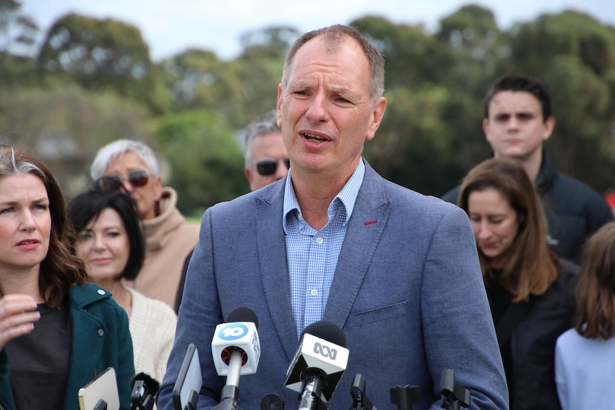 MP David Southwick speaks in a park at a press conference, with people behind him.