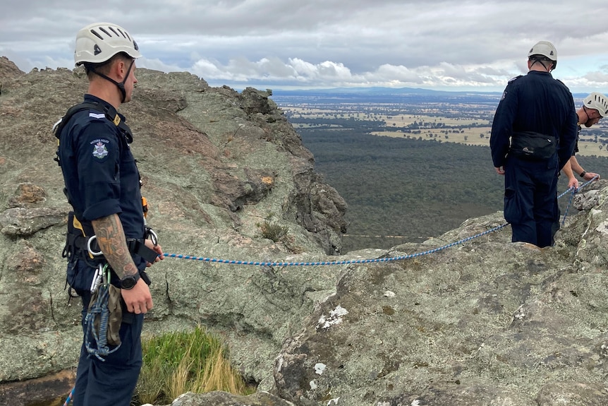 two men in navy uniform and a helmet attached together by ropes stand on the edge of a cliff overlooking the grampian.