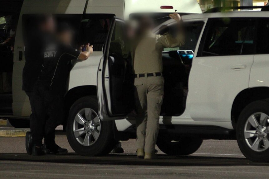 Three men stand around a white vehicle. Their faces are blurred.