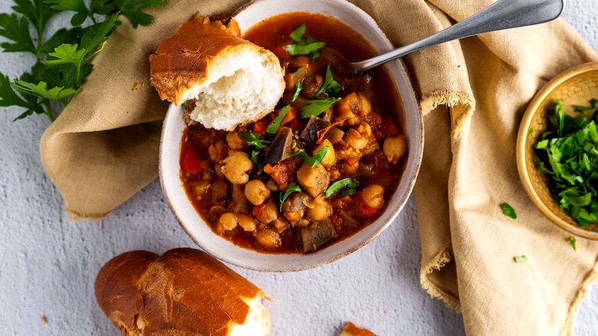 A bowl of chickpea and vegetable stew accompanied by fresh crusty bread and fresh herbs on the side.