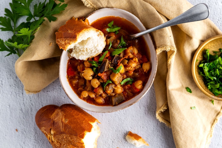 A bowl of chickpea and vegetable stew accompanied by fresh crusty bread and fresh herbs on the side.