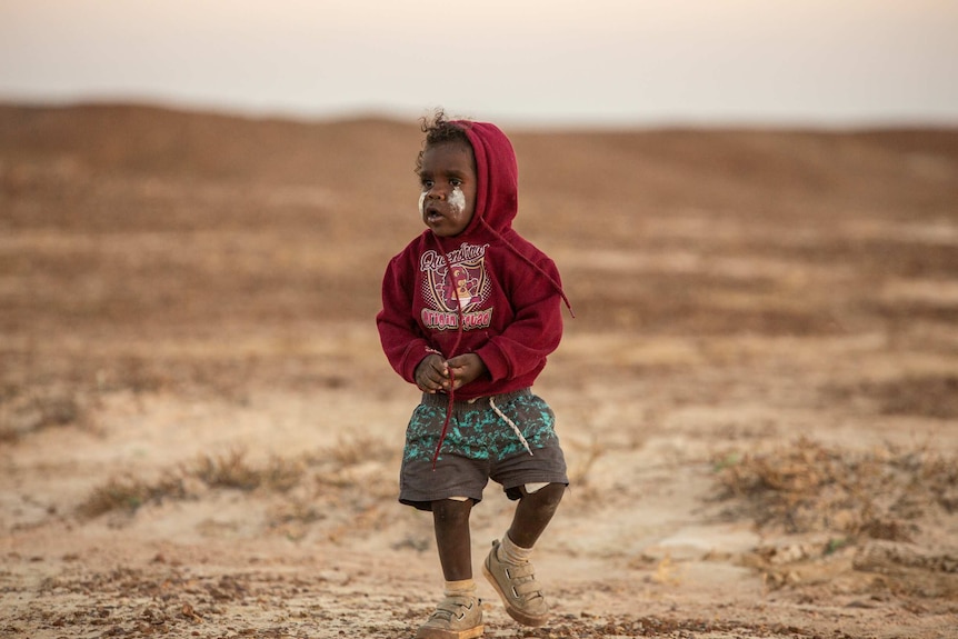 An indigenous toddler stands, with traditional paint on his face, on the Salt Flats of Burketown.