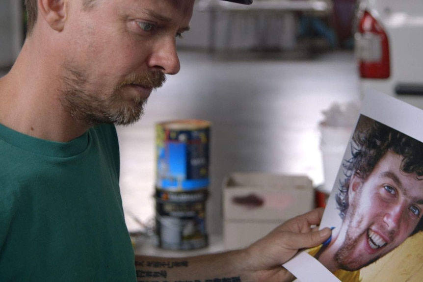 A man with a stubbly beard and a cap peers at printed out images of a young man with tousled black hair and a smile.