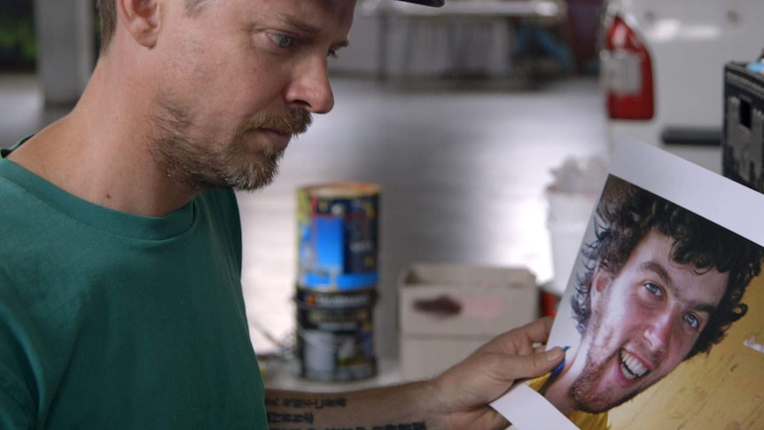 A man with a stubbly beard and a cap peers at printed out images of a young man with tousled black hair and a smile.