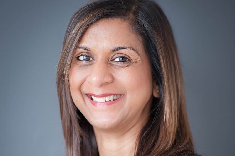 Nirvana Luckraj smiles as she poses for a professional portrait photograph in front of a grey background.