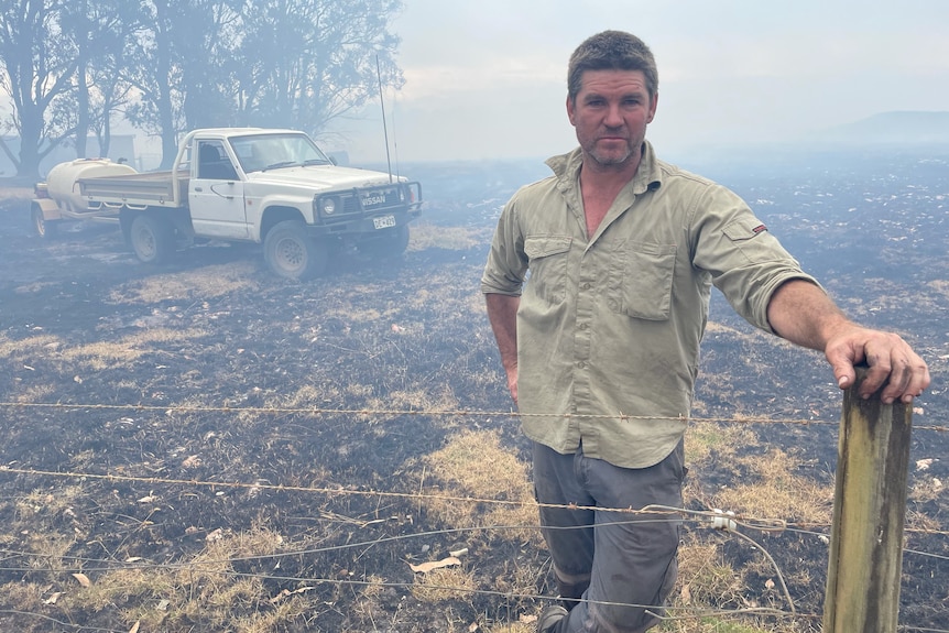 A man standing at a wire fence with a car in the background and lots of smoke and burnt floor and trees