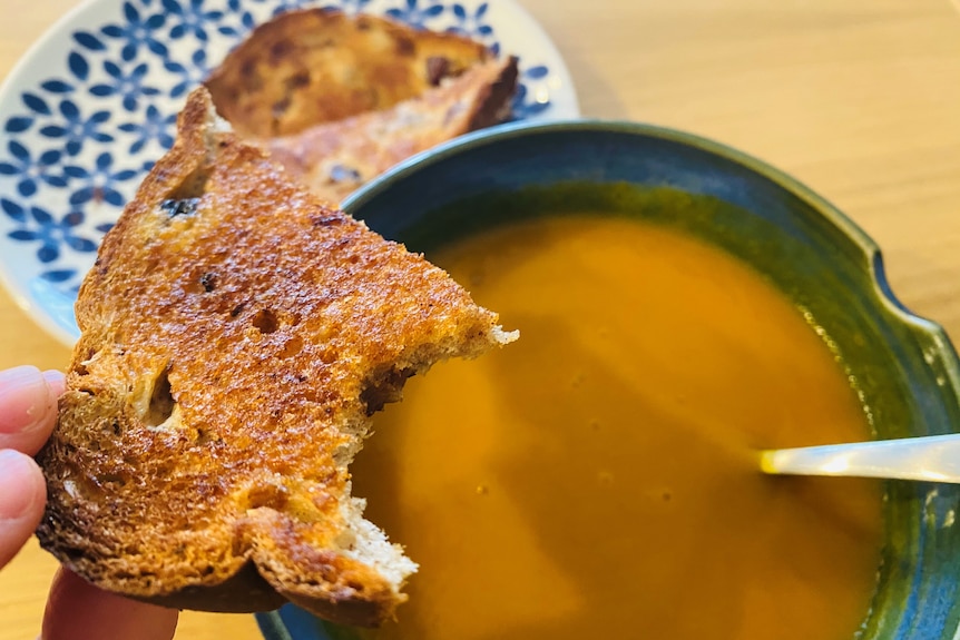 A slice of buttered raisin toast, held in front of a bowl of pumpkin soup, a proper lunch break.
