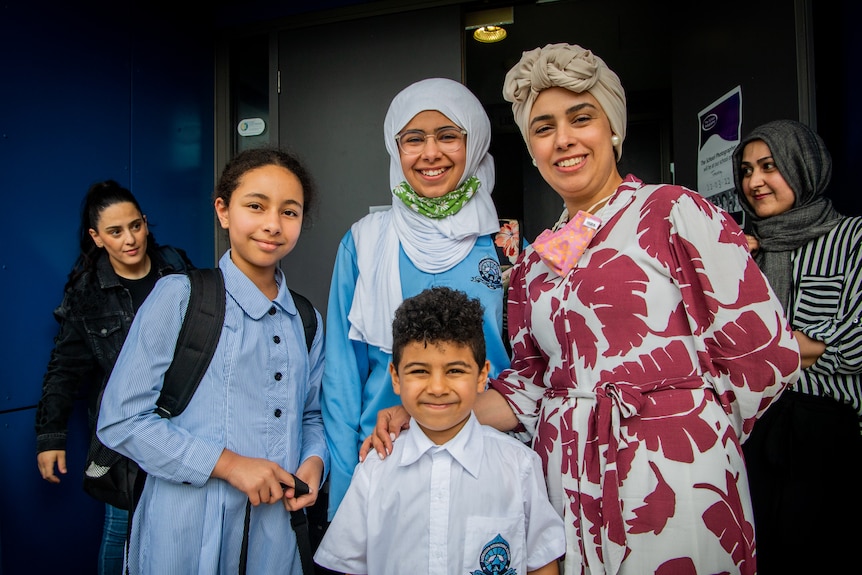 Nasha Mohammed wearing a bright long-sleeved dress, smiling with her three children outside a school.