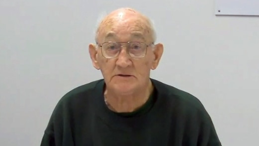An elderly man in glasses sits in front of a blank wall.