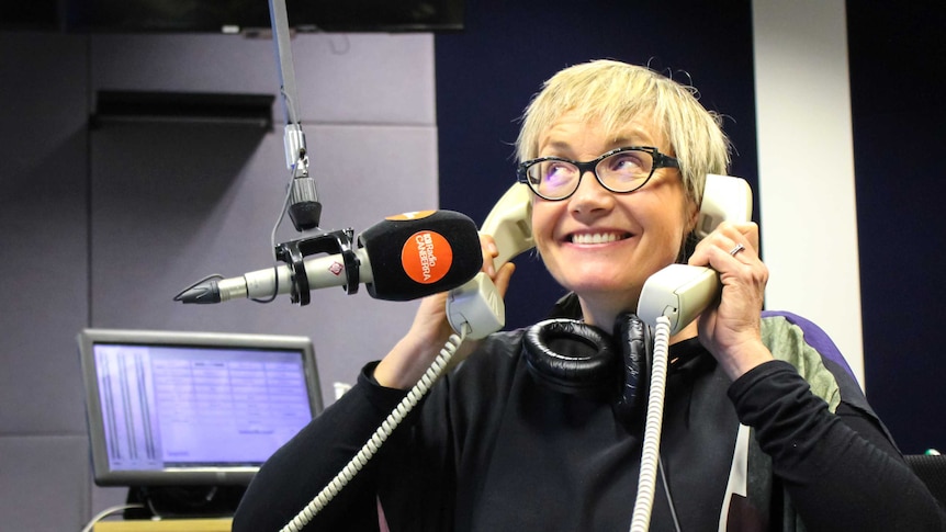 A woman is holding two desk phones to her ears in front of a radio studio microphone.