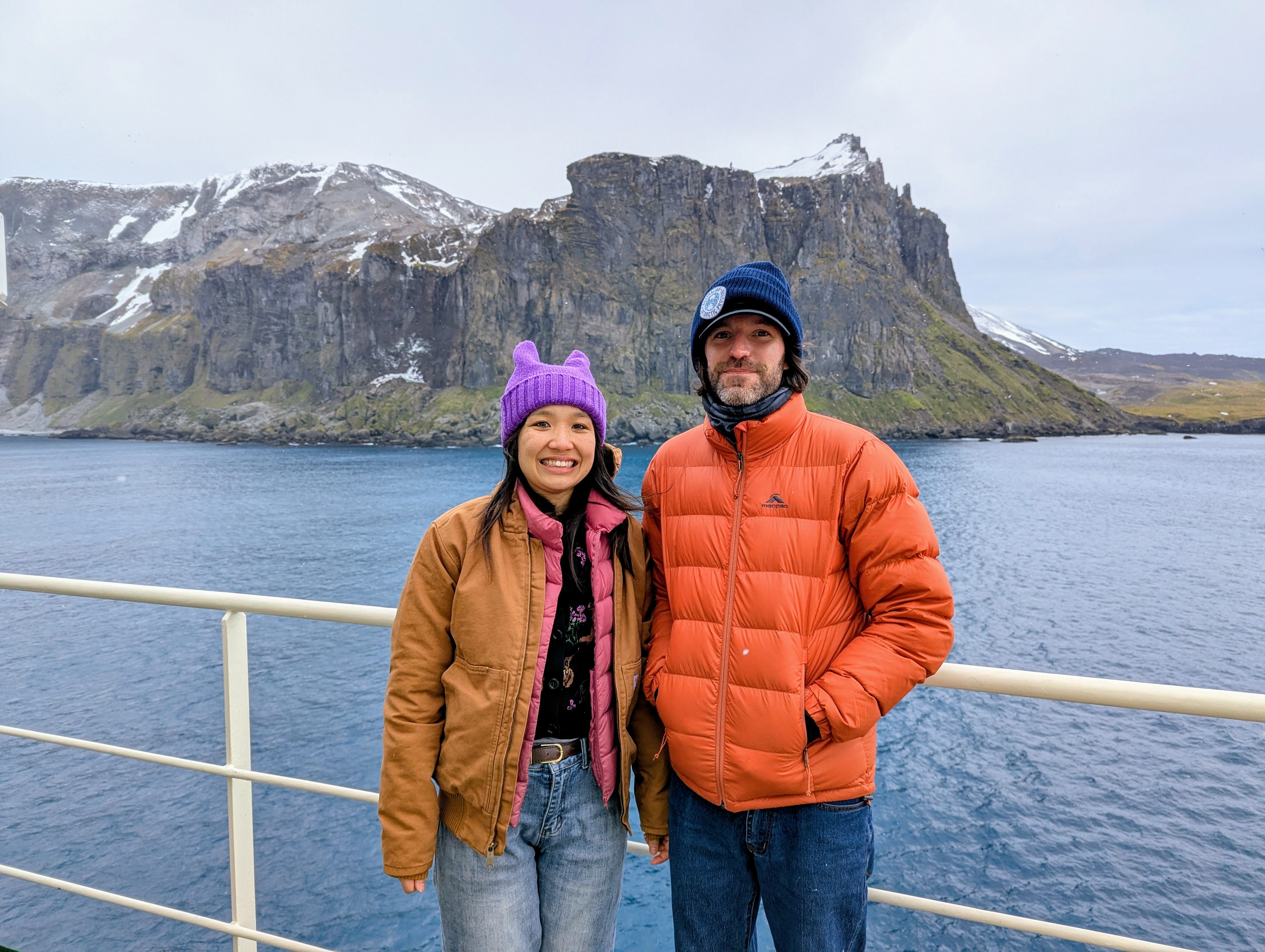 Winnie Yum and Nelson Athanasiou standing on a vessel in Antarctica, with snow-covered rocky terrain in background.