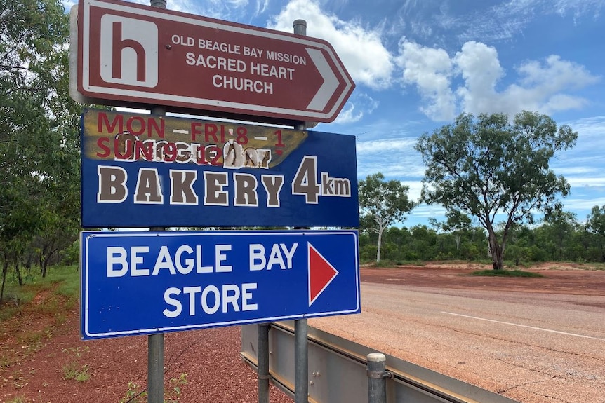Three roadside signs that say "Beagle Bay Store", "Bakery 4km" and "Old Beagle Bay Mission Sacred Heart Church".