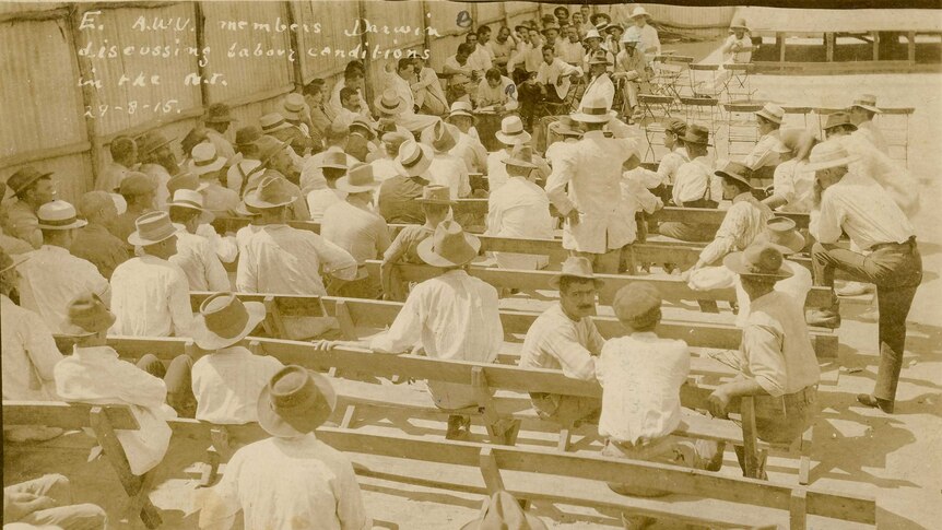 A group of men in white shirts and hats sit on wooden benches