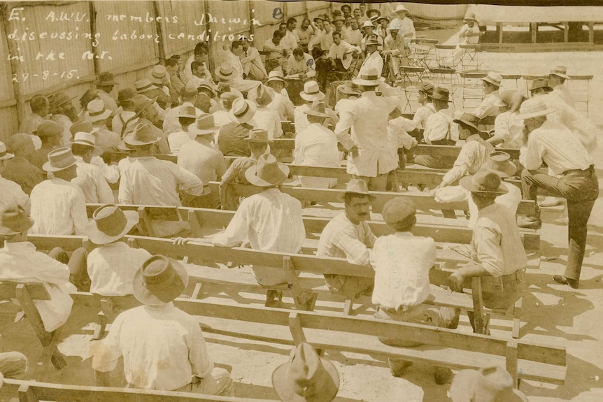 A group of men in white shirts and hats sit on wooden benches