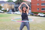 A woman smiles broadly while using a hula houp outside a block of units at a park in Sydney.