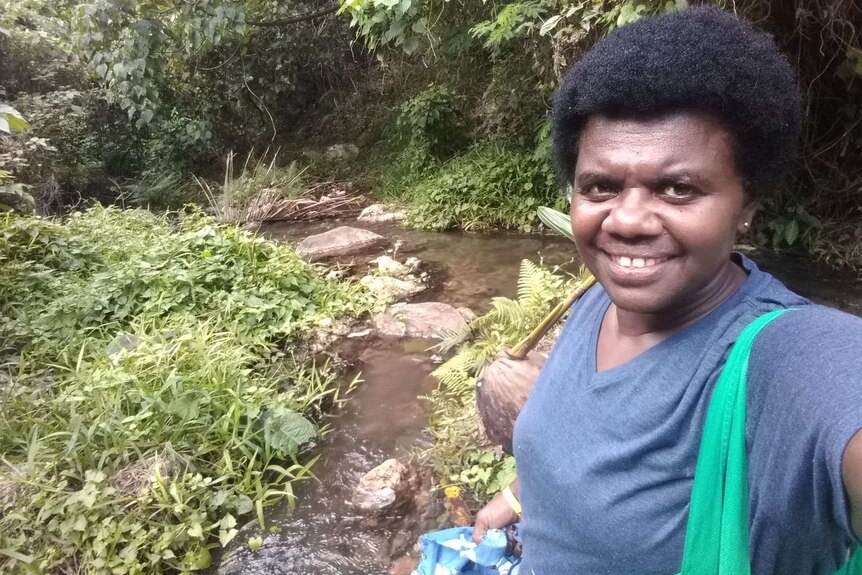 A woman with a bag on her shoulder smiles as she stands next to a stream