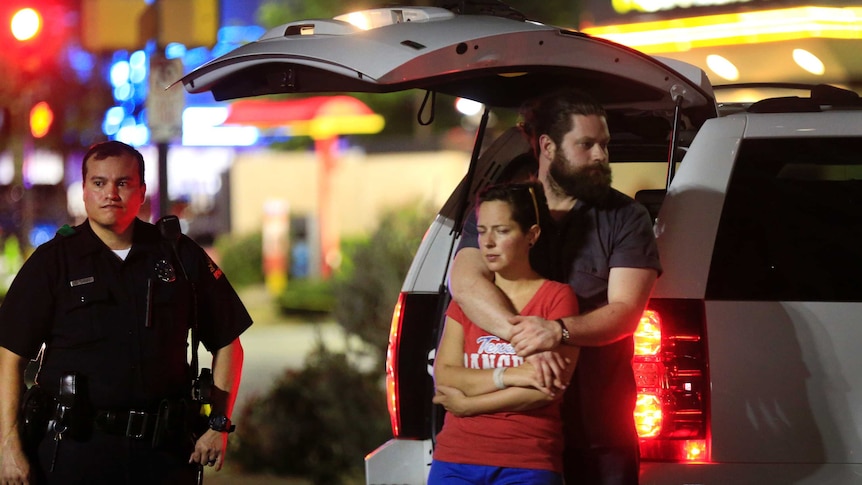 A police officer stands near a couple who are hugging and looking out at the scene.