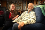 A very old man sitting in a soft chair with a woman crouched by his side, both smiling